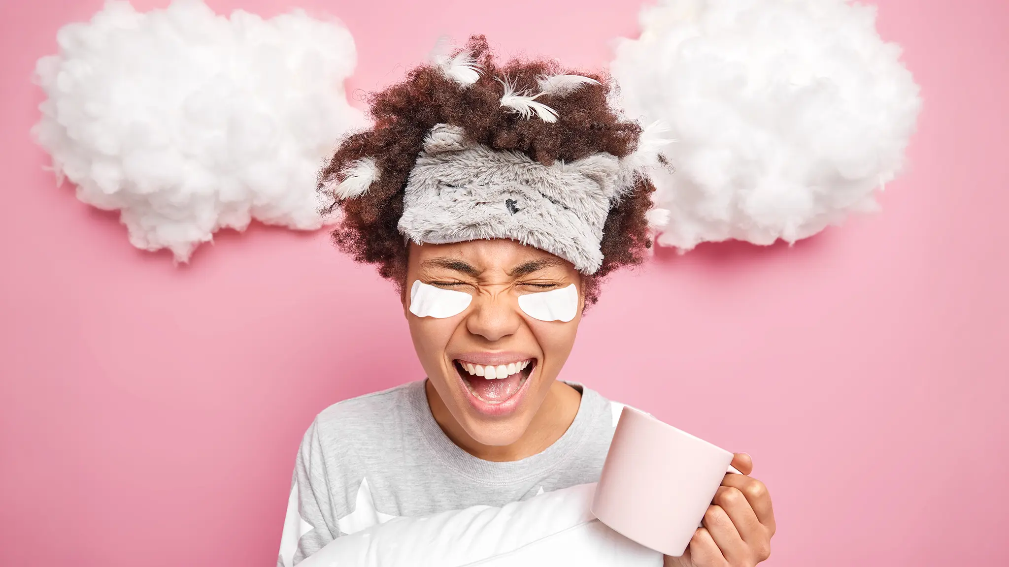 Junge Frau mit Schlafmaske, Augenpads und Kaffeetasse vor rosa Hintergrund – Symbol für Erschöpfung beim Bachelorarbeit Korrekturlesen