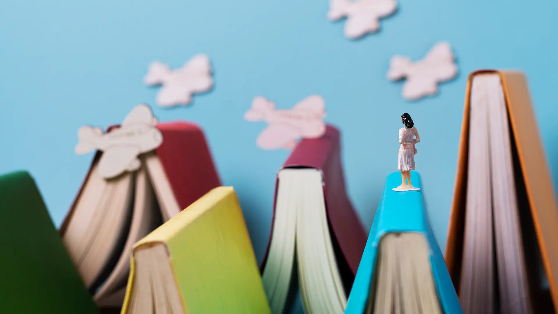 Texte übersetzen, lektorieren und gestalten – mit Kreativität und Präzision. Kleine Figur steht auf bunten Büchern vor Papierflugzeugen – symbolisch für kreatives Schreiben und Texte übersetzen.