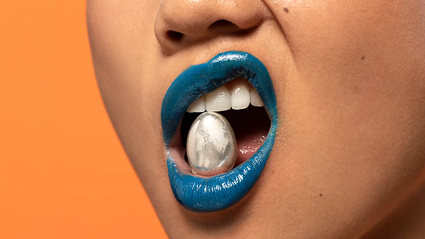 Audio- und Video-Transkription – Reden ist Silber, Text ist Gold Nahaufnahme von blau geschminkten Lippen mit einem silbern glänzenden Objekt im Mund; symbolische Darstellung für das Thema Audio- und Video-Transkription